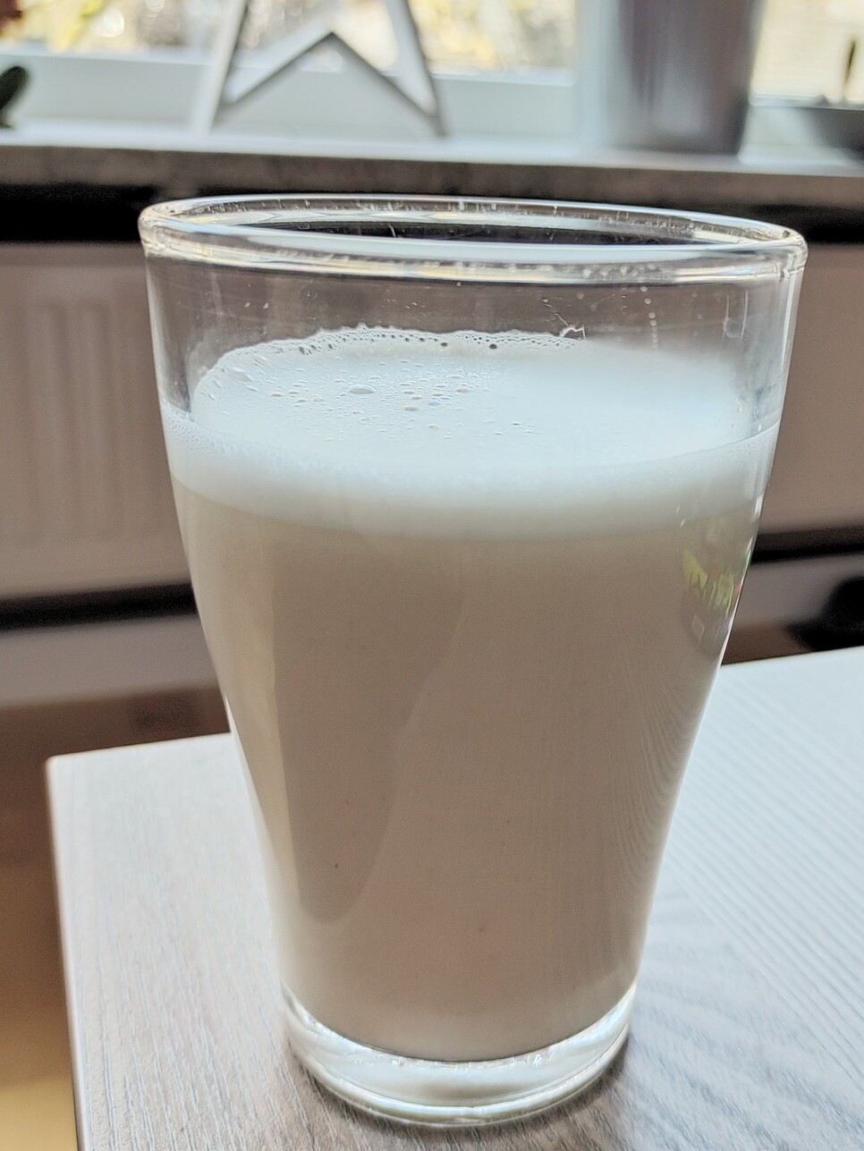 Hier ist ein Glas gefüllt mit leicht geschäumter Hafermilch auf einem Tisch vor einem Fenster zu sehen